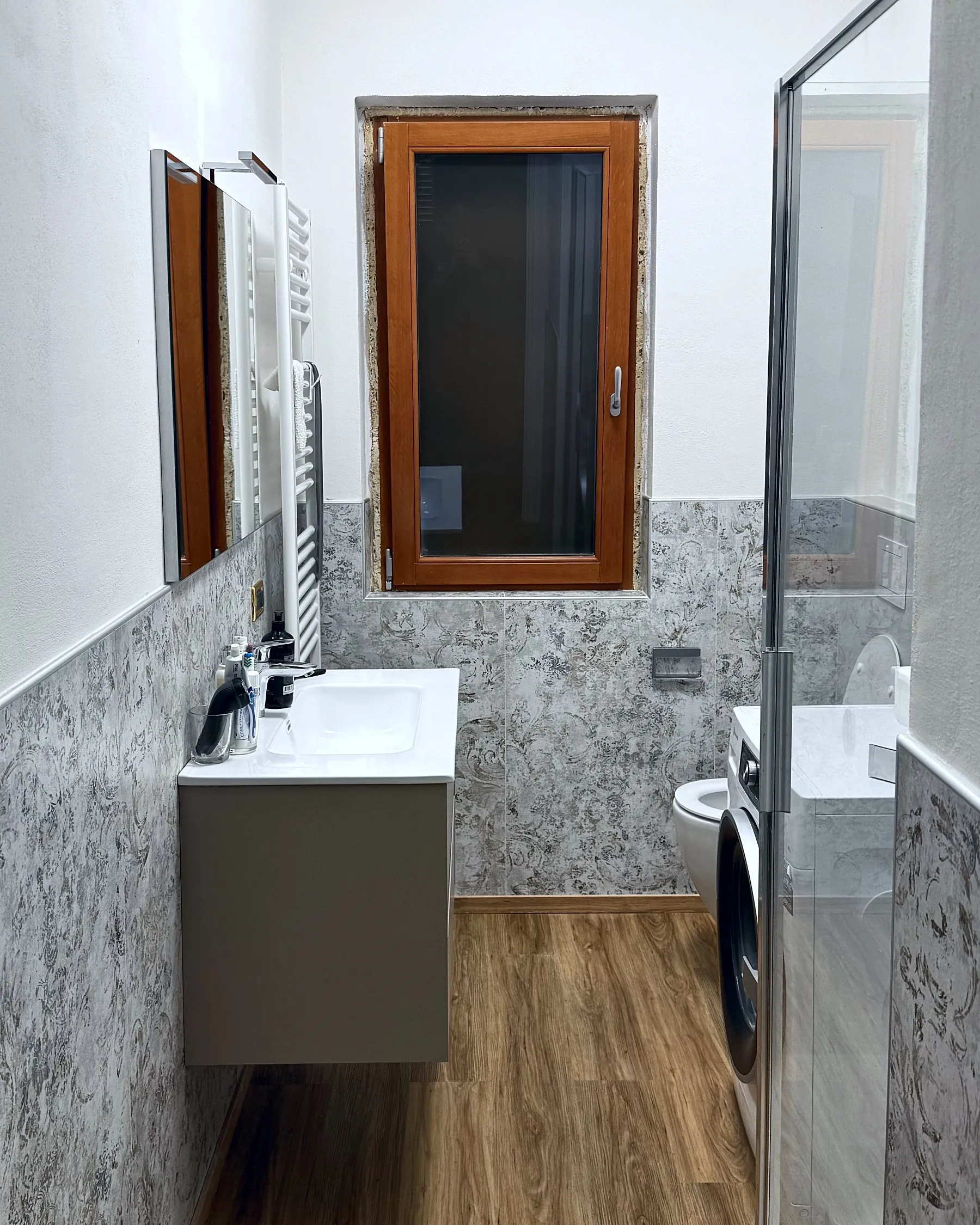 Bagno rifatto: finestra in legno, lavabo sospeso su mobile grigio, WC e lavatrice, rivestimento in pietra grigia